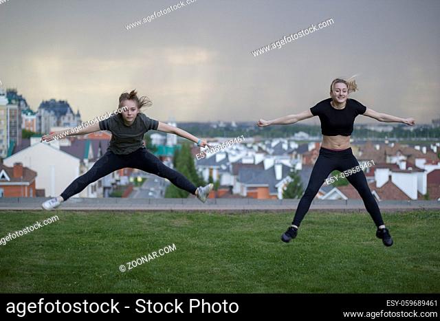 Two young female gymnasts doing acrobatics jumps with cityscape on background, horizontal