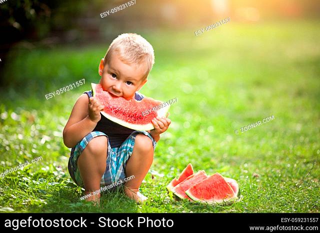 Cute toddler sitting outdoords and eating a slice of watermelon