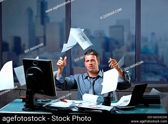 Exhausted businessman throwing documents into air sitting at office desk in overtime