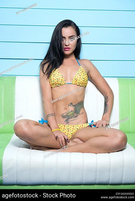 Beautiful seductive brunette woman wearing yellow bikini looking into the camera while sitting on the leather sofa over blue wooden wall background