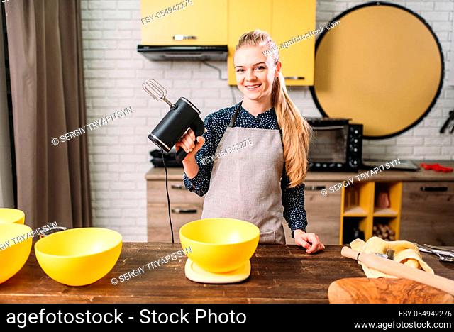 Young woman in apron holds mixer in hands against wooden table with bowls for dough. Sweet cake cooking preparation. Kitchen on background