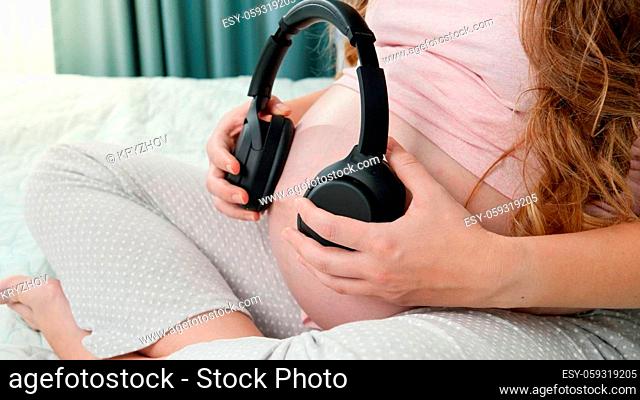 Closeup of pregnant woman sitting on bed and putting headphones on her big belly. Unborn baby listening to classic music while being in mothers abdomen