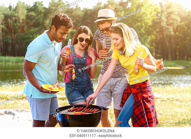 Happy young friends are making barbecue in the nature. They are standing near grill and smiling. Men and women are drinking beer