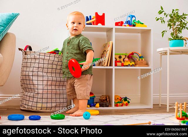 The little blond boy takes toys out of the bag. He stands in the playroom with a toy and looks around