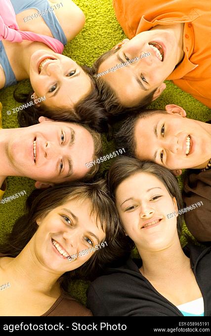 Group of 6 teenagers lying on the floor head next to head. They're looking at camera and smiling