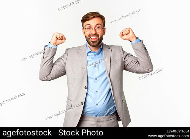 Portrait of happy businessman with beard, wearing grey suit and glasses, raising hands up in hooray gesture, saying yes and rejoicing
