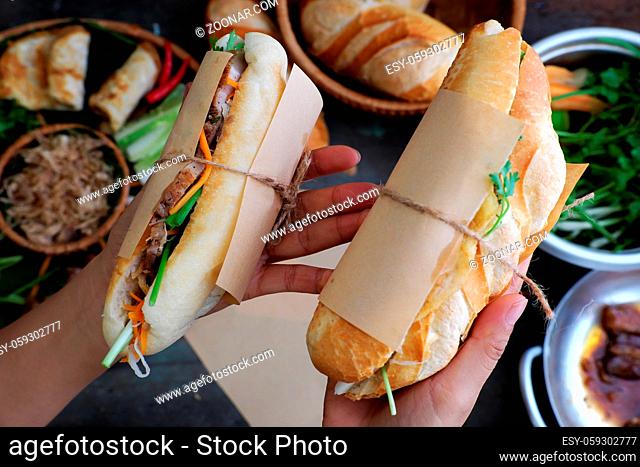 Woman hand make banh mi thit or Vietnamese bread, famous street food from raw material: pork, ham, pate, egg and fresh herbs as scallions, coriander, carrot