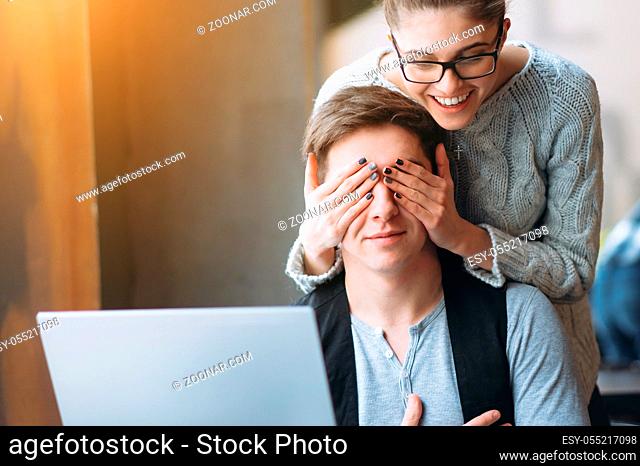 Young man typing on laptop in cafe. His girlfriend making him surprise and closing eyes