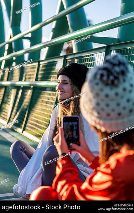 Woman wearing knit hat taking photo of smiling friend through mobile phone while sitting on bridge
