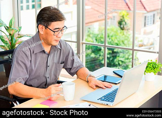 Asian executive senior businessman sitting on desk office with his office, the confident middle aged handsome man using laptop computer at workplace home office...