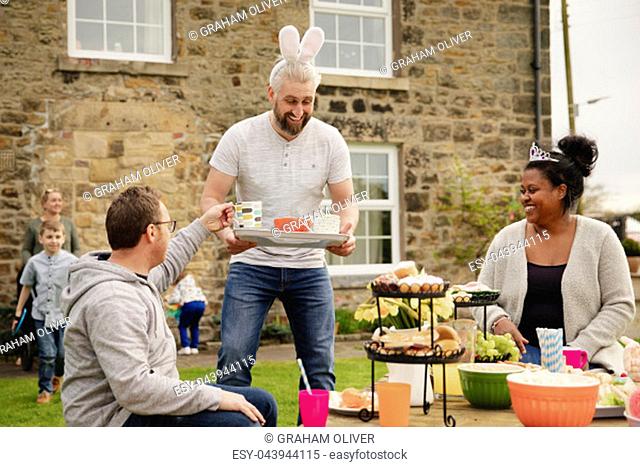 Mid adult man carrying a tray with cups of tea. He is handing out refreshments while wearing rabbit ears at a easter garden party