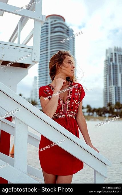 Beautiful woman wearing red dress leaning on wooden structure of lifeguard hut at beach