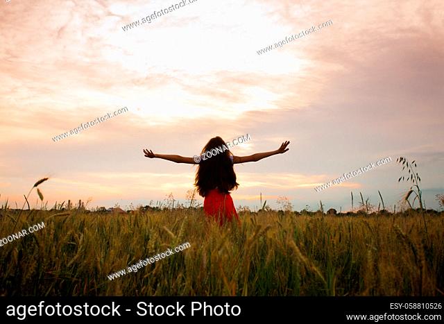 Young girl in red skirts is standing with arms outstretched in a field. A beautiful girl is standing with her back to the camera