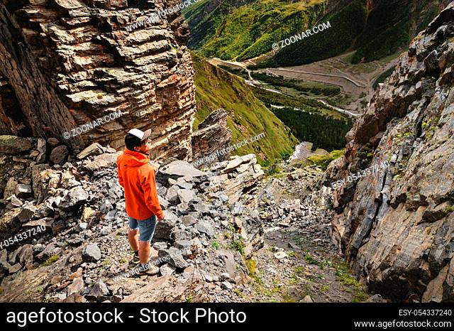 Portrait of a male hiker in shorts and a cap from the back is standing on high rocks at the edge of a cliff against the background of a green valley with a road...