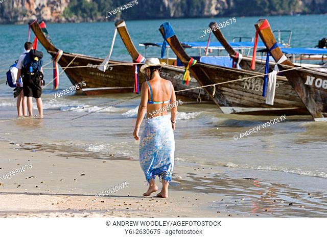 Woman in sun hat bikini and sarong walks beach by longtail boats Ao Nang Thailand