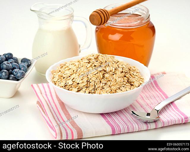 Morning breakfast, raw oatmeal flakes in a ceramic plate, milk in a decanter and honey in a jar on a white table, close up