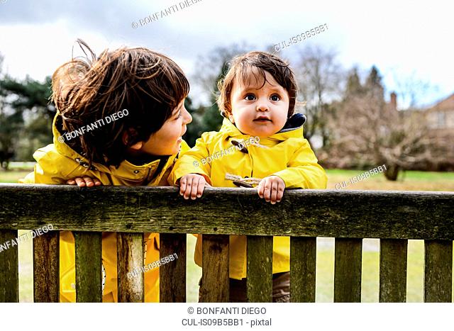 Portrait of baby boy and big brother in yellow anoraks on park bench