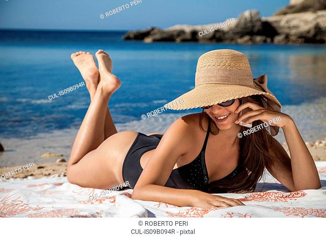 Young woman wearing sunhat chatting on smartphone whilst sunbathing on beach, Villasimius, Sardinia, Italy