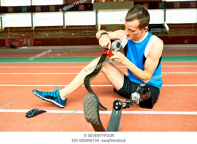 Full length portrait of young amputee sportsman sitting on floor in modern gym fixing prosthetic foo, copy space