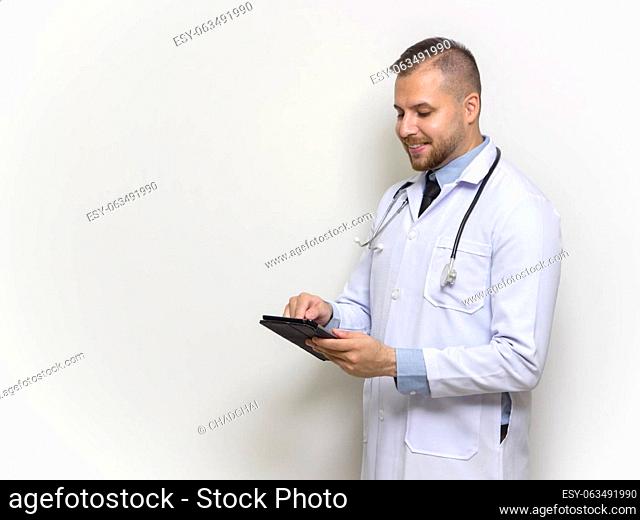 Young caucasian doctor with short hair, mustache and beard. Dressed in white gown and stethoscope typing on tablet computer