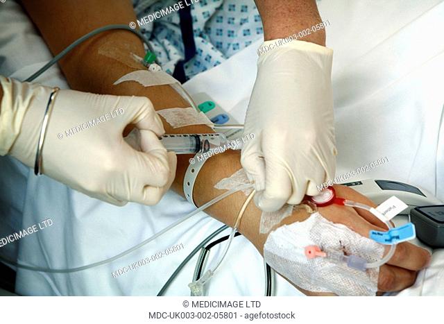 The gloved hand of a nurse injects fluid into a catheter which is attached to the arm of a patient