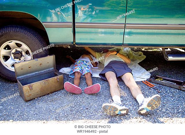 Man and a boy lying under a vehicle with a toolbox, doing repairs