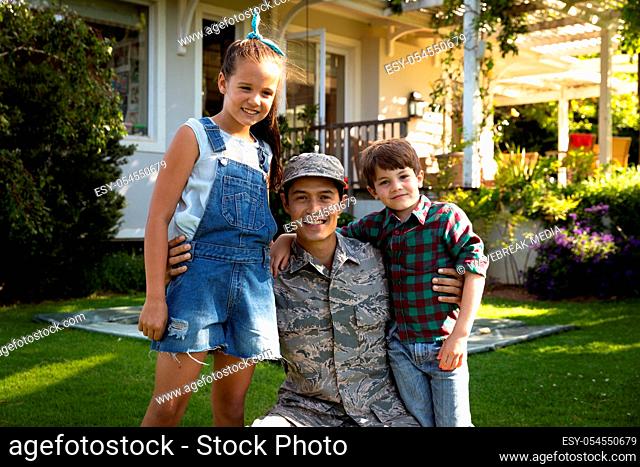 Portrait of a young adult mixed race male soldier in the garden outside his home, kneeling with his arms around his young son and daughter