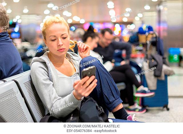 Casual blond young woman using her cell phone while waiting to board a plane at the departure gates. Wireless network hotspot enabling people to access internet...