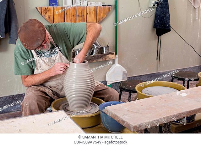 Potter wearing flat cap sitting at pottery wheel shaping clay vase