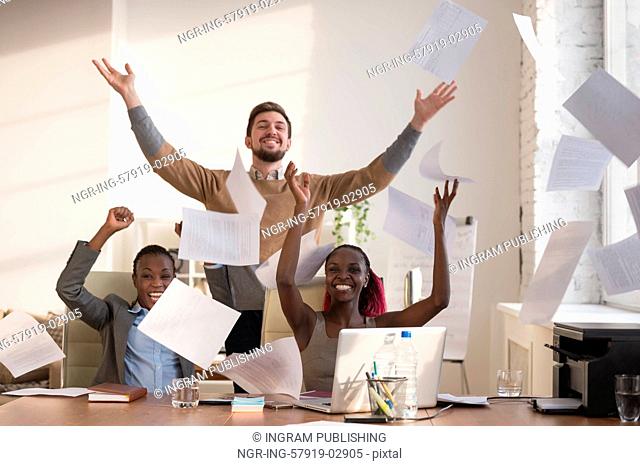 Business people excited happy smile, throwing up papers, documents fly in air, businesspeople sitting at office desk hold hands arms up, success team concept
