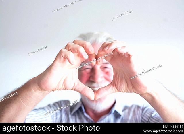 close up and portrait of mature man and senior doing an heart with his fingers and hand in front of the camera - happy pensioner having fun enjoying - white...