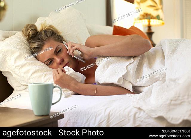 Young woman with tissue in her bed talking to doctor on smartphone, coffee mug sitting on bedside table