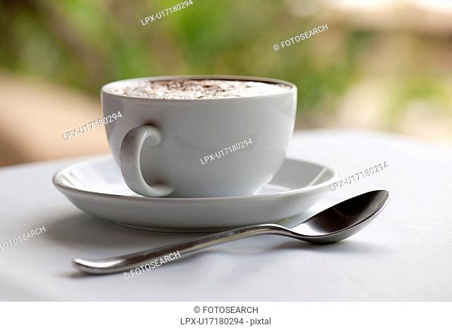 Cofffee cup, saucer and teaspoon, filled with steamed milk and sprinkled with spice, on a white table