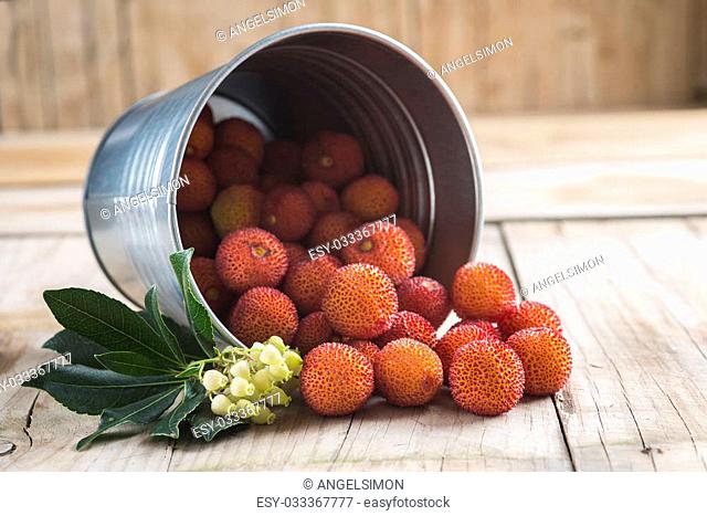 Bucket with ripe arbutus unedo fruits, leaves and flowers on a wooden background