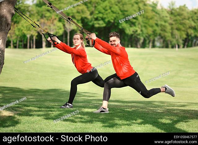 Group of sport people exercising with suspension trainer in green park. Happy man and woman in red jackets looking at camera