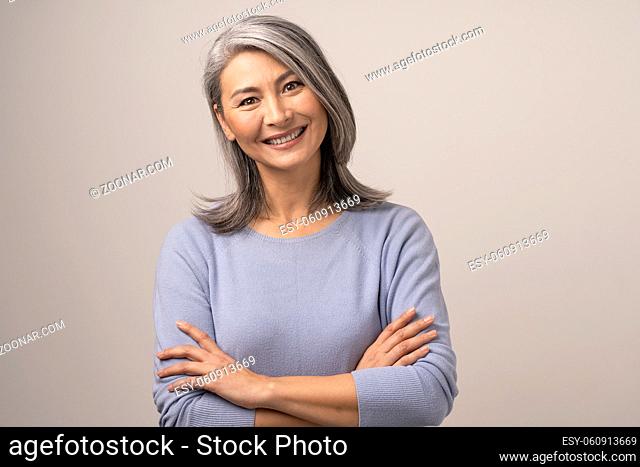 Happy Woman With Broad Smile And Crossed Arms. Cute Asian Grey-Haired Woman Broadly Smiles And Crosses Her Arms Posing At Studio On White Background