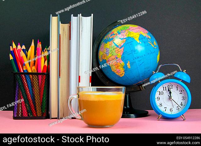 Coffe is in the foreground and behind him are a stack of books, a globe, a clock and a glass of pencils