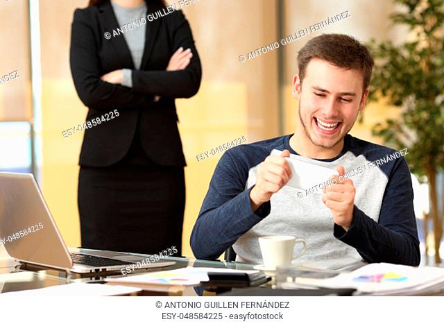 Lazy employee playing games with his smartphone sitting in a desktop while his angry boss is watching at office