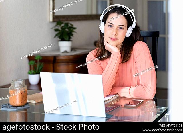 Beautiful businesswoman with hand on chin listening music through headphones at home