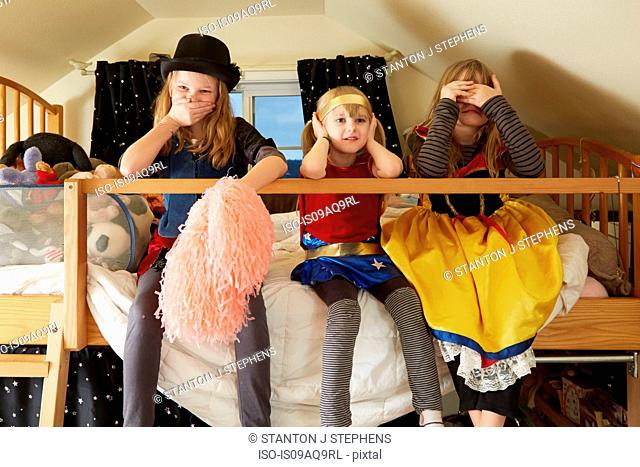 Three sisters, sitting on bed, wearing fancy dress costumes, covering mouth, ears and eyes