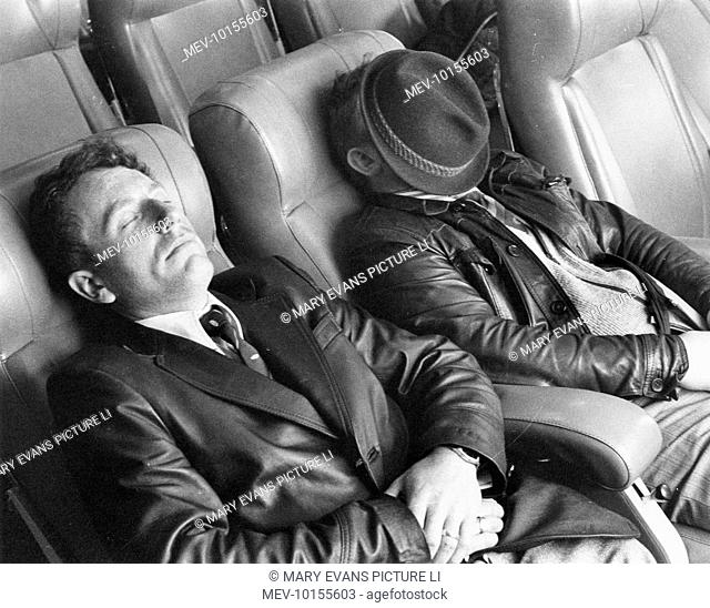 Two men in leather jackets, one of them with his hat over his face, take a nap on a cross Channnel ferry, perhaps having had too much booze on their cruise