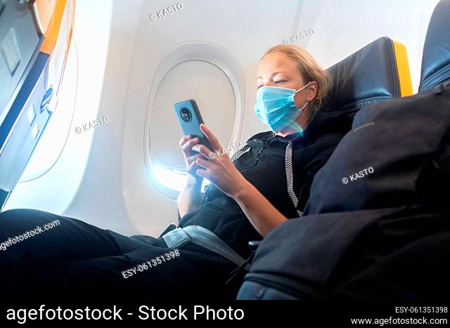 A young woman wearing face mask, using smart phone while traveling on airplane. New normal travel after covid-19 pandemic concept