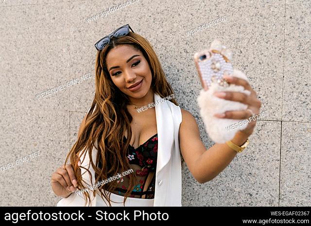 Fashionable woman taking selfie through smart phone while standing in front of wall