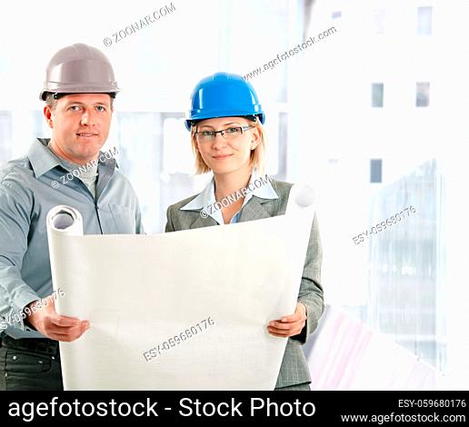 Creative engineer coworkers working on plan in hardhat, smiling at camera