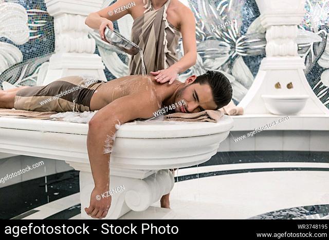 Relaxed young man lying down on hot marble bed during traditional Turkish bath at luxury spa and wellness center