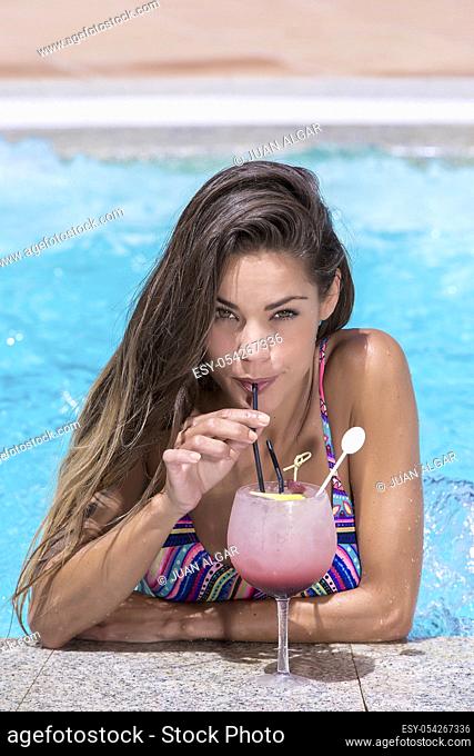 Beautiful female in colorful swimsuit drinking cocktail through straw leaning on spa pool edge and looking in camera