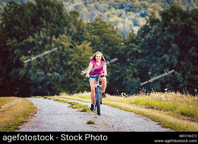 Girl on bicycle cycling down a dirt path in summer, woods in the background