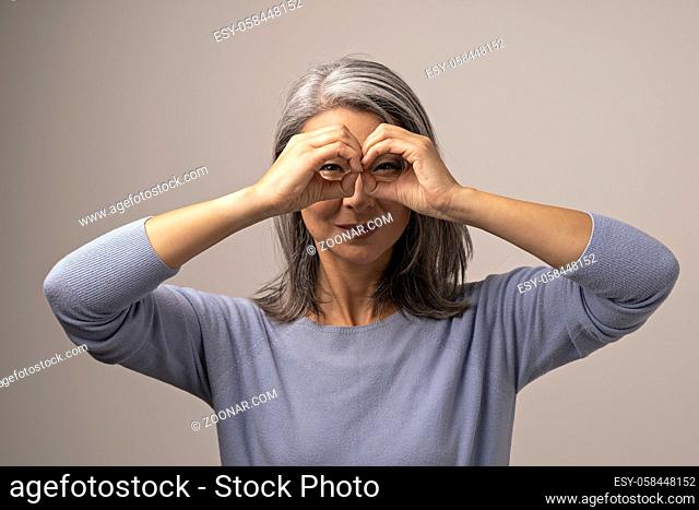 Beautiful Grey-Haired Woman Squints Her Eyes While Looking Through Her Hands As If They Are Glasses. Asian Woman Squinting Her Eyes While Doong Ok Gesture Like...