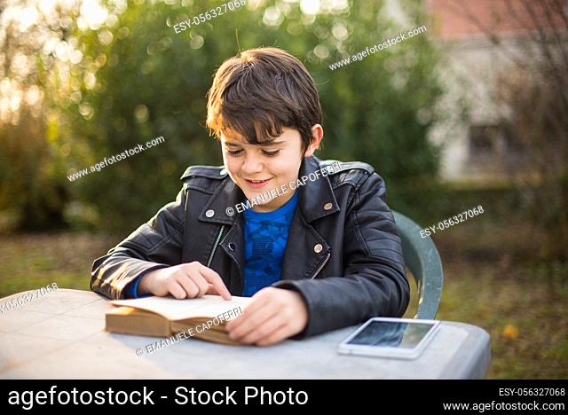 young boy reads book, smiling, sitting in garden, tablet on table