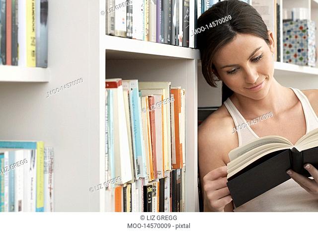 Young woman reading book leaning against bookshelf